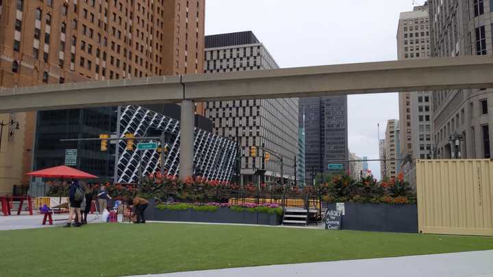 Urban plaza with a stage and flowers in front of a downtown scene and an elevated people mover track. 