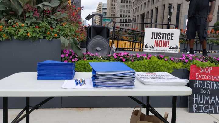 Stacks of blue folders and signs that say “No evictions!” on a white folding table in front of a stage, yard sign that says “Justice now! DetroitEvictionDefense.org. 313-530-0216.” and a handwritten protest sign that says “Stop evictions. Housing is a human right.” 