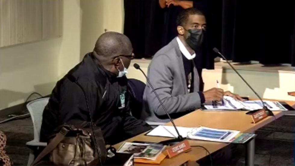 QuanTez Pressley (with darker skin, black hair, face mask, grey suit) speaking to someone off-screen. Cedric Banks (with darker skin, bald head, glasses, face mask, brown suit) looks at Pressley. 