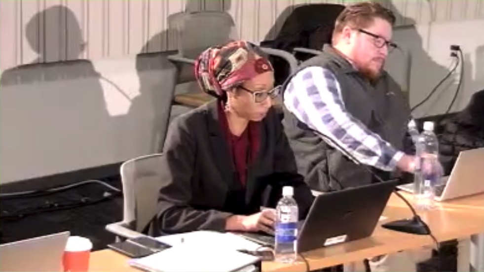 A woman with darker skin and wearing a colorful head wrap, glasses, black coat is speaking and looking at her laptop screen. 