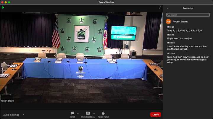 Online meeting software showing a video feed of an empty meeting room with tables in a U-shaped arrangement. At the front of the room is a “City of Detroit” banner flanked by United States and Detroit flags, next to a television screen showing a green graphic. 