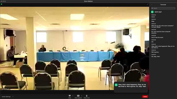 Online meeting application showing a video feed of a white room with seats arranged for an audience to face a long table draped in blue cloth. 