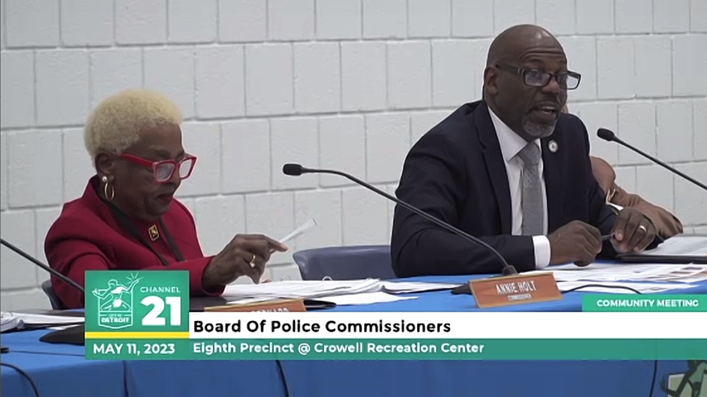 Black man speaking into microphone on blue table with many pieces of paper and a nameplate that says, “Bryan Ferguson. Commissioner.” He is wearing glasses and a suit. He is sitting to the right of a Black woman wearing red glasses and red attire. 