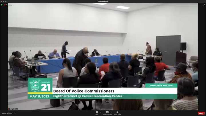 Audience of at least 20 people, seated, facing six people sitting at a wide conference table draped in blue, with more empty seats. Two people are walking to the table. The room is painted white without windows. The graphic at the bottom says, “Channel 21. Board of Police Commissioners. Community Meeting. May 11, 2023. Eighth Precinct @ Crowell Recreation Center.” 