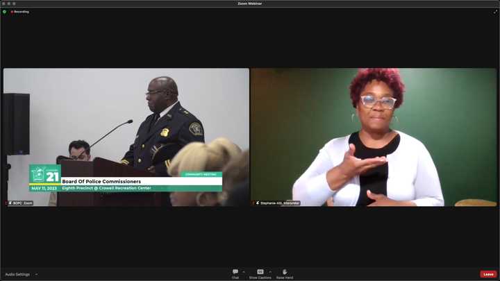 Person speaking at podium. They are wearing glasses and a black, formal Detroit Police uniform, and they have dark skin and a bald head. Sign language interpreter Stephanie is wearing glasses, earrings, white blouse and black shirt; they have brown skin and brown hair. 